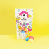 Colorful space-themed bath bomb packaging on a bright yellow background. The playful design features cartoon UFOs and stars, conveying a fun and cheerful vibe.