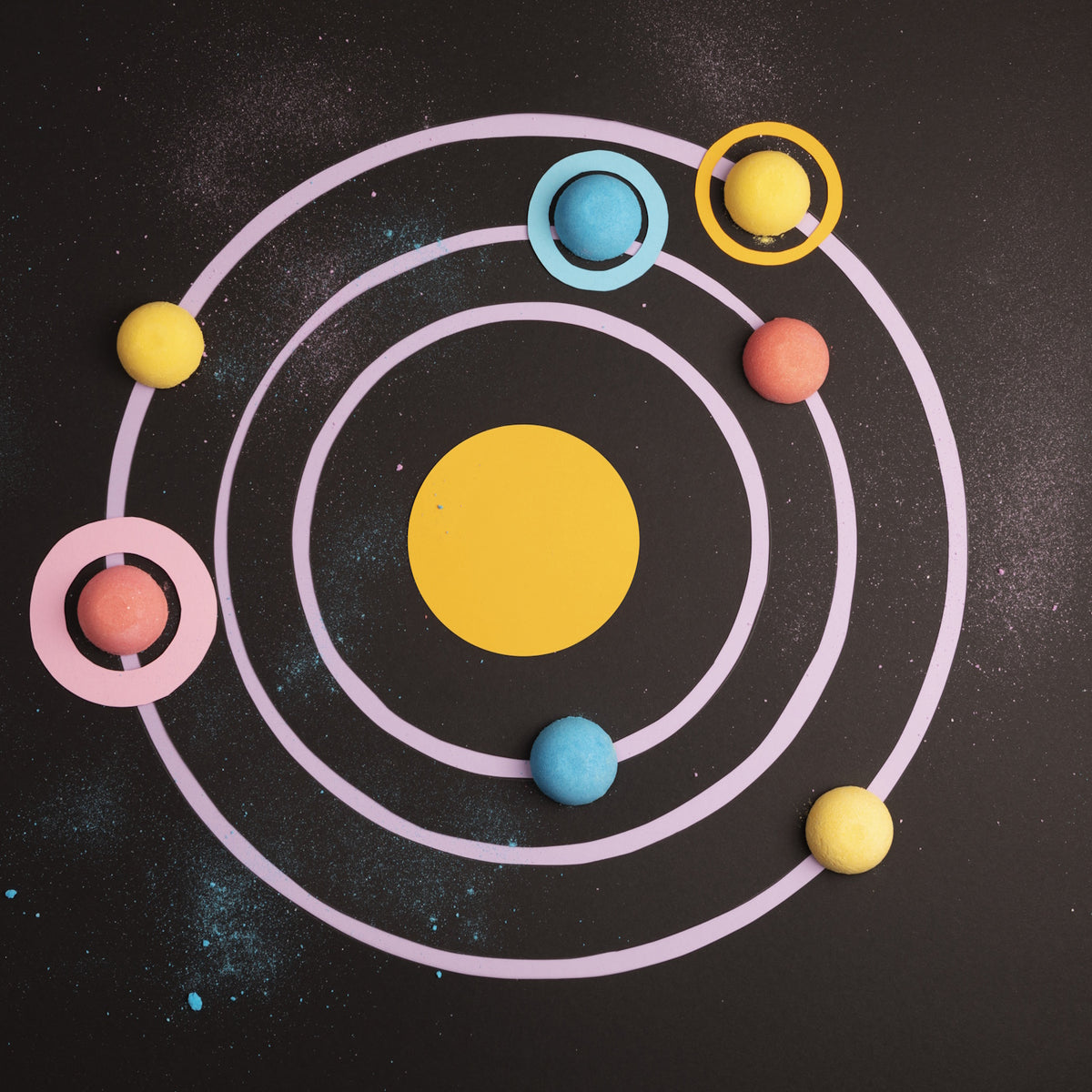 Colorful, candy-like planets orbit a yellow circle on a dark, cosmic background with pastel rings and scattered specks, evoking a playful solar system.