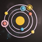 Colorful, candy-like planets orbit a yellow circle on a dark, cosmic background with pastel rings and scattered specks, evoking a playful solar system.