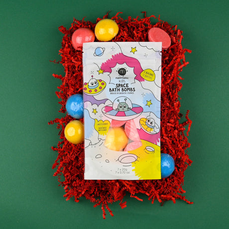 A colorful package of kids' space-themed bath bombs is placed on red shredded paper. The playful design features cartoon aliens and planets. The background is green.