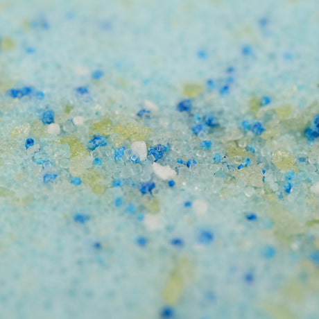 Close-up of multicolored bath salt crystals on a blue background. The tiny grains sparkle in shades of blue, white, and green, creating a textured appearance.