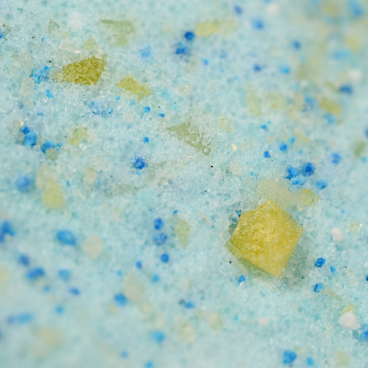 Close-up of colorful bath salt granules, featuring a mix of blue, white, and yellow particles. The texture is coarse and scattered.