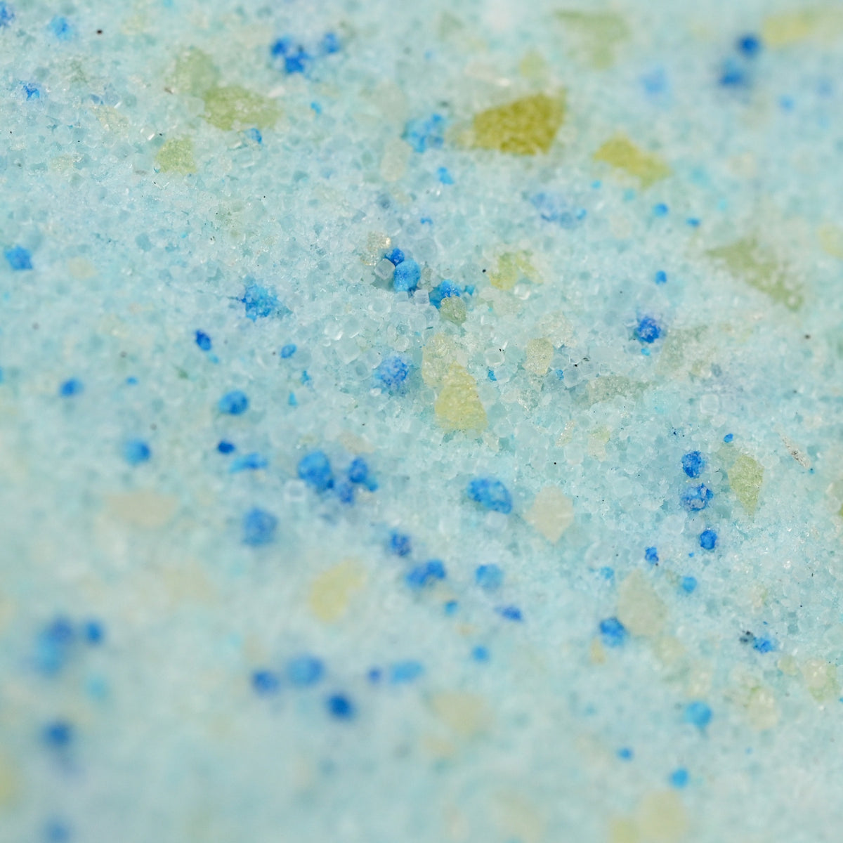 Close-up of a textured surface with granules in shades of light blue, green, and yellow, resembling crystals or bath salts, evoking a refreshing feel.
