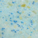 Close-up of a textured surface with granules in shades of light blue, green, and yellow, resembling crystals or bath salts, evoking a refreshing feel.