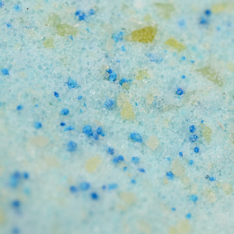 Close-up of a textured surface with granules in shades of light blue, green, and yellow, resembling crystals or bath salts, evoking a refreshing feel.