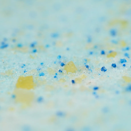 Close-up of a granular mixture of blue and yellow particles on a light blue background, resembling bath salt. The texture is coarse and colorful.