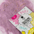 Packet of crackling bath salts labeled "Pop!" and "Snap!" on a playful design. Rose pink salts are scattered underneath, adding a whimsical, fun vibe.