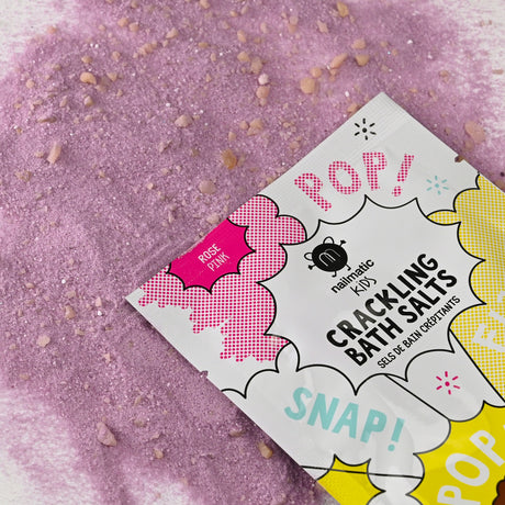Packet of crackling bath salts labeled "Pop!" and "Snap!" on a playful design. Rose pink salts are scattered underneath, adding a whimsical, fun vibe.