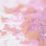 Close-up of pink and orange crystalline bath salt scattered on a white surface. The salt crystals vary in size, creating a playful and delicate atmosphere.