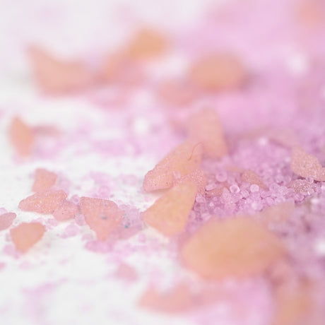 Close-up of pink and orange crystalline bath salt scattered on a white surface. The salt crystals vary in size, creating a playful and delicate atmosphere.