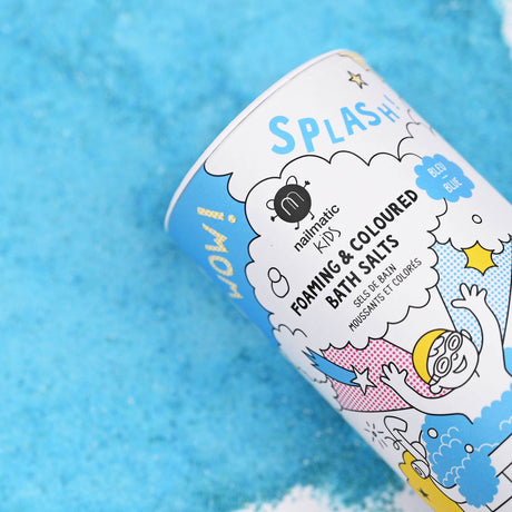 Packaging for Nailmatic Kids foaming bath salts, with colorful illustrations of a superhero and bubbles, lies on a bright blue salt surface. Playful and fun.