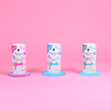 Three colorful bath salt containers stand on a pink background. Each has playful cartoon graphics, and vibrant salts spill out below: purple, blue, and green.