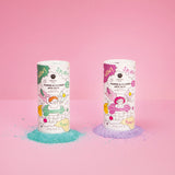 Two cylindrical containers of foaming bath salts stand on a pink background. One has green illustrations with spilled blue powder; the other has pink illustrations with spilled purple powder. Both feature playful graphics, creating a fun, vibrant mood.