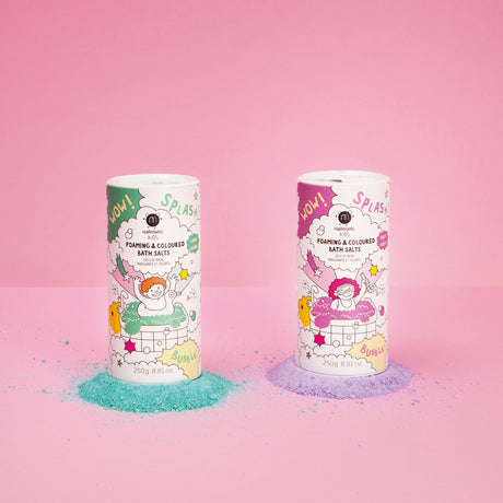 Two cylindrical containers of foaming bath salts stand on a pink background. One has green illustrations with spilled blue powder; the other has pink illustrations with spilled purple powder. Both feature playful graphics, creating a fun, vibrant mood.