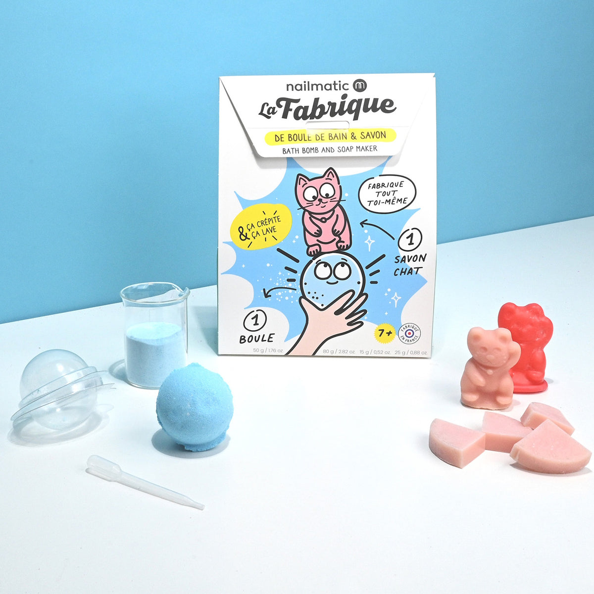 A DIY bath bomb and soap maker kit features a colorful box with a cute cat illustration, surrounded by pink soaps, a blue bath bomb, and measuring tools.