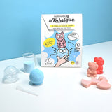 A DIY bath bomb and soap maker kit features a colorful box with a cute cat illustration, surrounded by pink soaps, a blue bath bomb, and measuring tools.