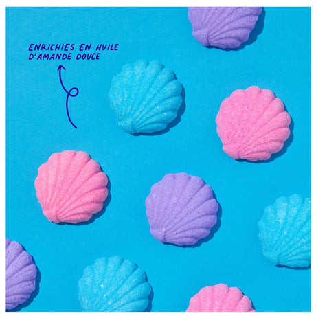 Colorful shell-shaped bath bombs in pink, blue, and purple are arranged on a bright blue background. Text reads, "ENRICHIES EN HUILE D'AMANDE DOUCE."