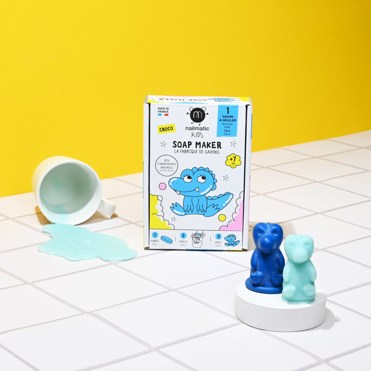 Soap making kit on white tiled surface with spilled blue liquid from a cup. Two cute animal-shaped soaps beside the box against a bright yellow backdrop.