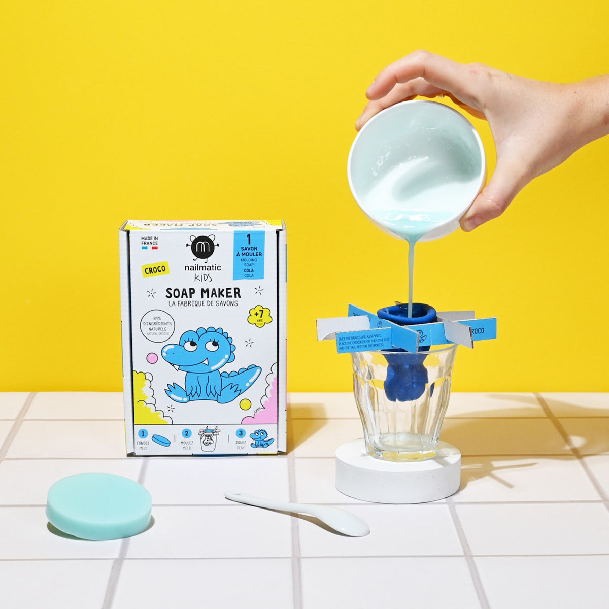 A box with a cartoon crocodile design labeled "Nailmatic Kids Soap Maker" sits next to a hand pouring blue liquid into a mold over a glass. The background is bright yellow.