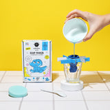 A box with a cartoon crocodile design labeled "Nailmatic Kids Soap Maker" sits next to a hand pouring blue liquid into a mold over a glass. The background is bright yellow.