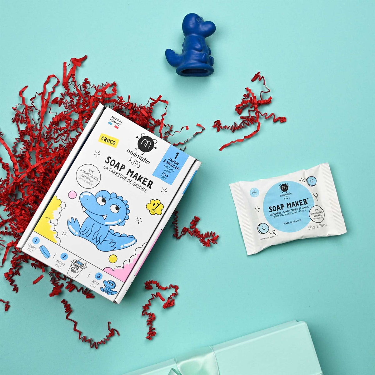 A kids' soap-making kit with a blue crocodile character on the packaging lies on a cyan background, surrounded by red paper shreds and a soap mold.