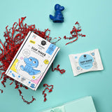 A kids' soap-making kit with a blue crocodile character on the packaging lies on a cyan background, surrounded by red paper shreds and a soap mold.