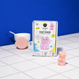 Box of Nailmatic Kids Soap Maker Kit with pink cat illustration on a tiled surface. Nearby, a pink cat-shaped soap and a bowl with a round soap. Playful and colorful.