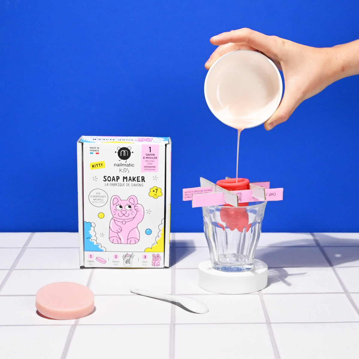 A hand pours pink liquid into a soap mold over a glass. Nearby, a colorful kids' soap maker kit box with a cartoon cat and pink soap disc are visible.