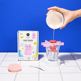 A hand pours pink liquid into a soap mold over a glass. Nearby, a colorful kids' soap maker kit box with a cartoon cat and pink soap disc are visible.
