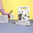 A hand draws a black tattoo of a rabbit on the wrist using a nailmatic Tattoo Pen. A tattoo kit with colorful designs is on a stone surface.