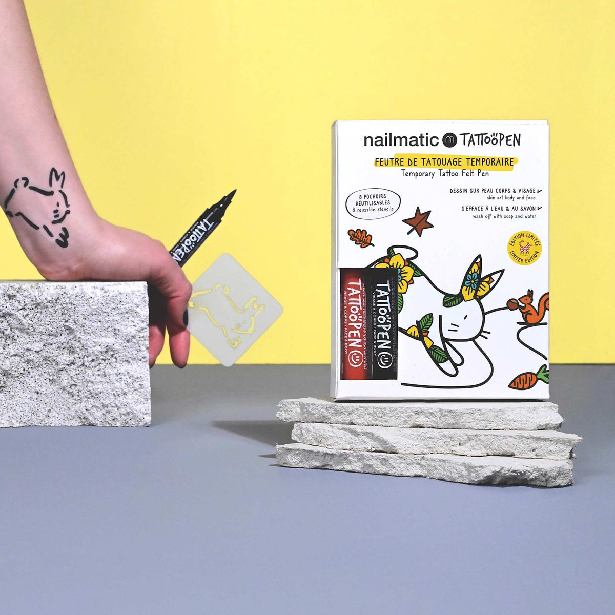 A hand draws a black tattoo of a rabbit on the wrist using a nailmatic Tattoo Pen. A tattoo kit with colorful designs is on a stone surface.