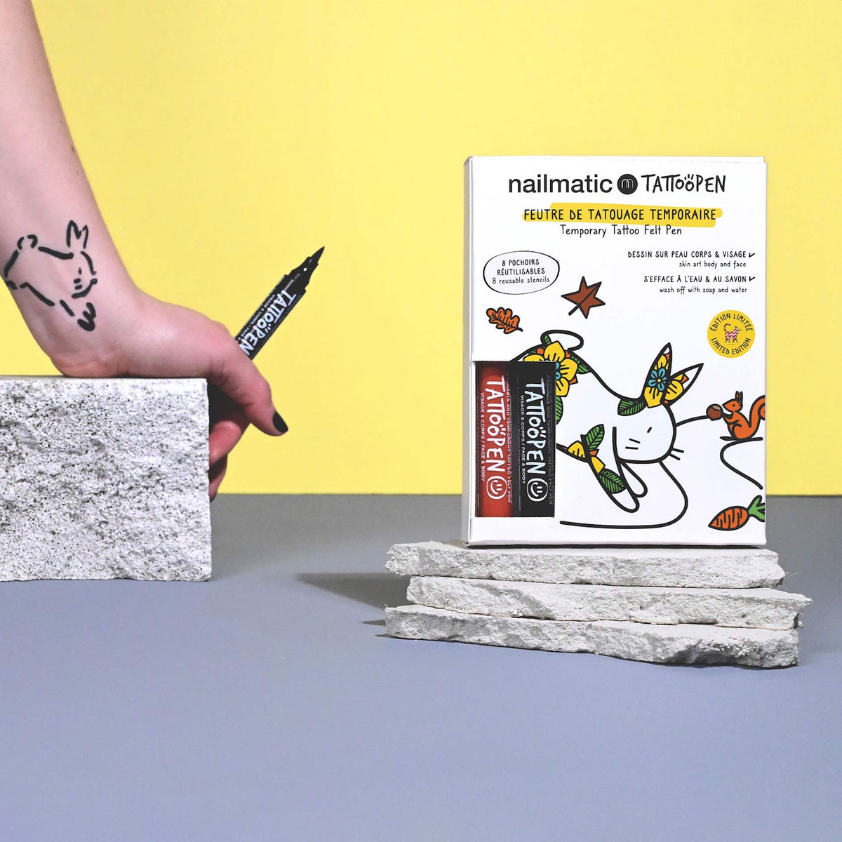 A hand holding a Tattoo Pen is drawing a simple tattoo on an arm. A colorful Tattoo Pen box is placed on stacked stones against a yellow background.