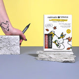 A hand holding a Tattoo Pen is drawing a simple tattoo on an arm. A colorful Tattoo Pen box is placed on stacked stones against a yellow background.