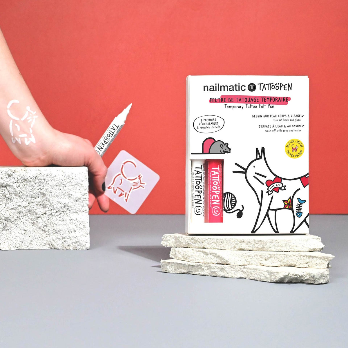 A hand holds a white tattoo pen drawing a cat on skin with a stencil. A box of temporary tattoo pens, featuring cat designs, stands nearby on stone blocks.