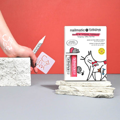 A hand holds a white tattoo pen drawing a cat on skin with a stencil. A box of temporary tattoo pens, featuring cat designs, stands nearby on stone blocks.