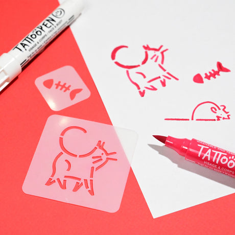 Red and pink stencils of a cat and fish are used on white paper with a pink tattoo marker and a white tattoo pen on a vibrant red background.