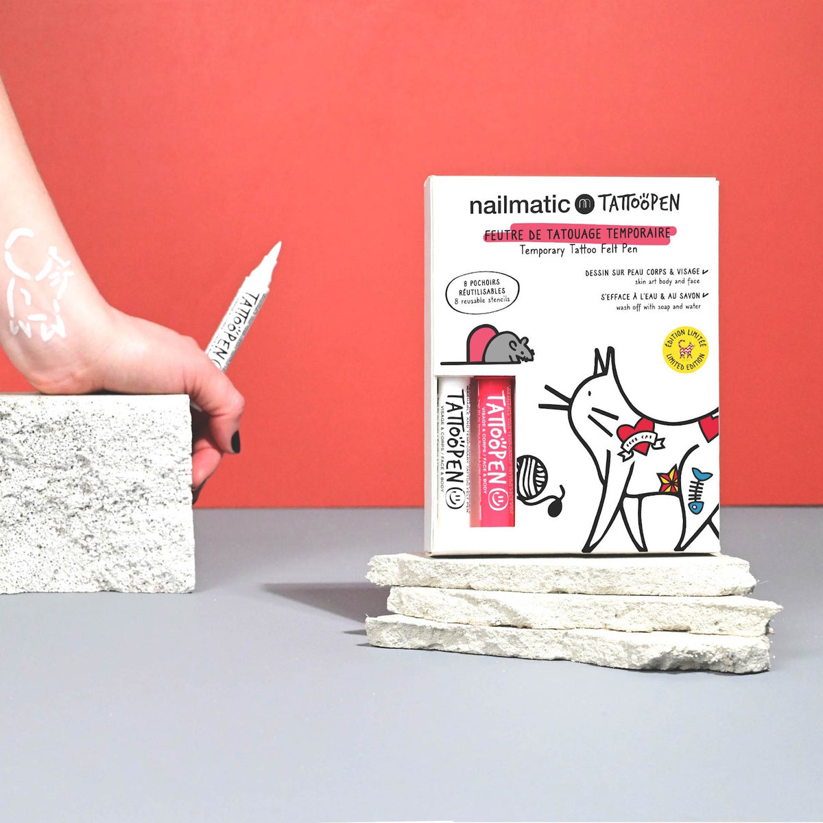 Hand using white pen to draw on skin, next to boxed tattoo pen set. Background is red and gray, creating a playful, creative vibe.