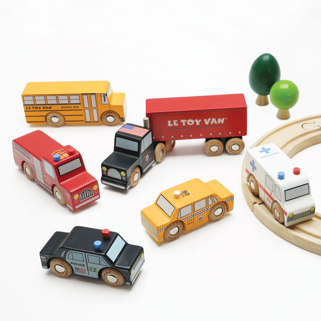 Wooden toy vehicles including a red semi-truck, yellow school bus, black police car, white ambulance, red fire truck, and yellow taxi, all with colorful details.