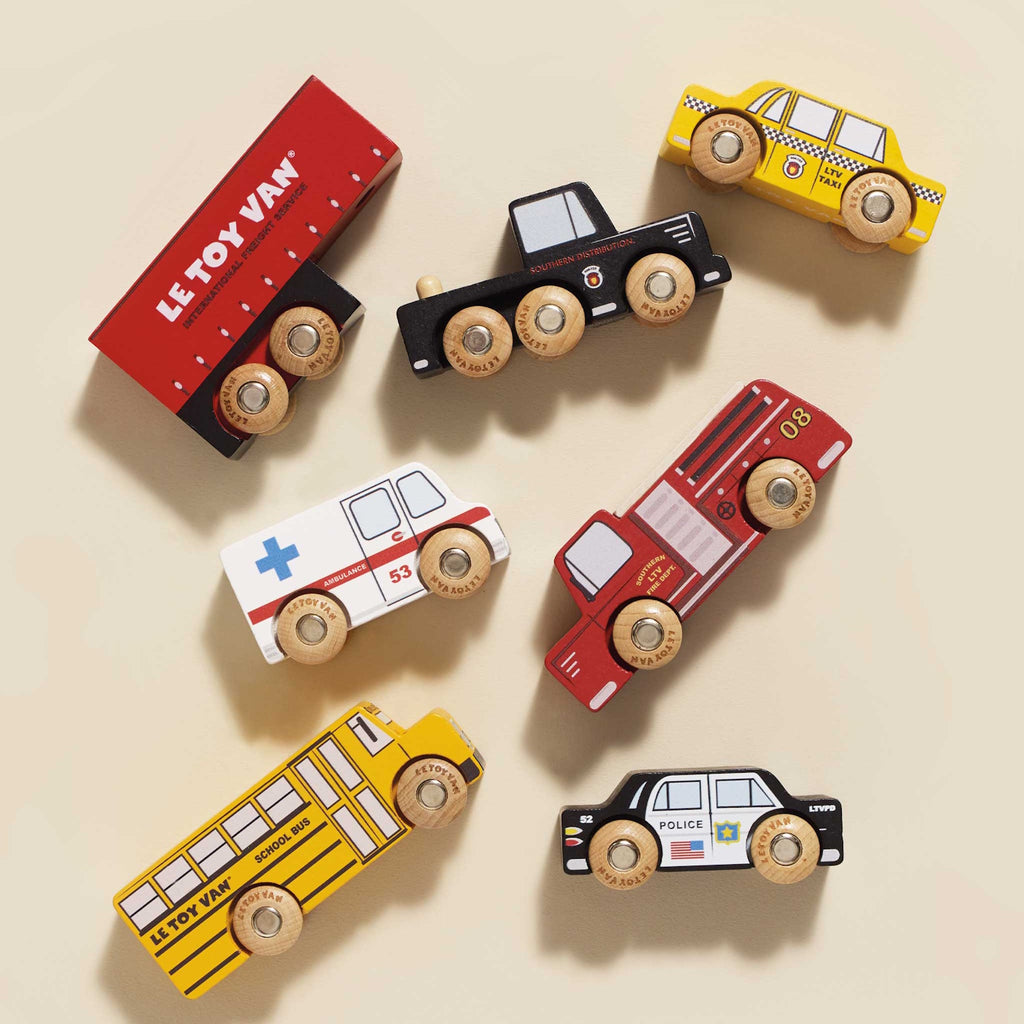 Wooden toy vehicles including a red semi-truck, yellow school bus, black police car, white ambulance, red fire truck, and yellow taxi, all with colorful details.
