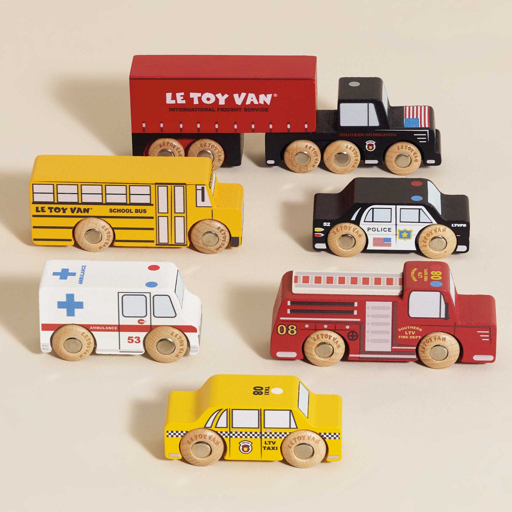 Wooden toy vehicles including a red semi-truck, yellow school bus, black police car, white ambulance, red fire truck, and yellow taxi, all with colorful details.