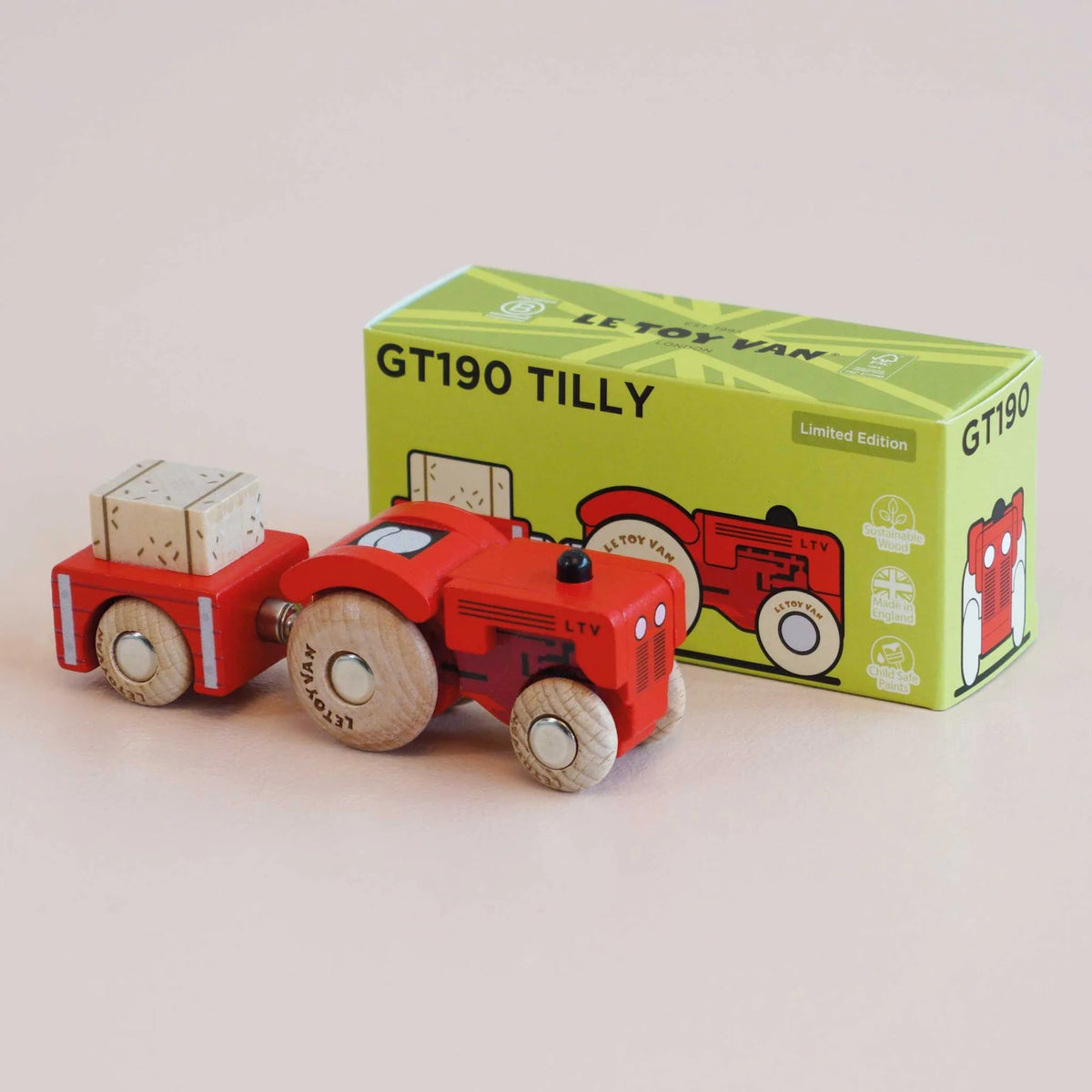 Red wooden toy tractor with trailer and hay bale positioned next to its green packaging box in a front three-quarter view.