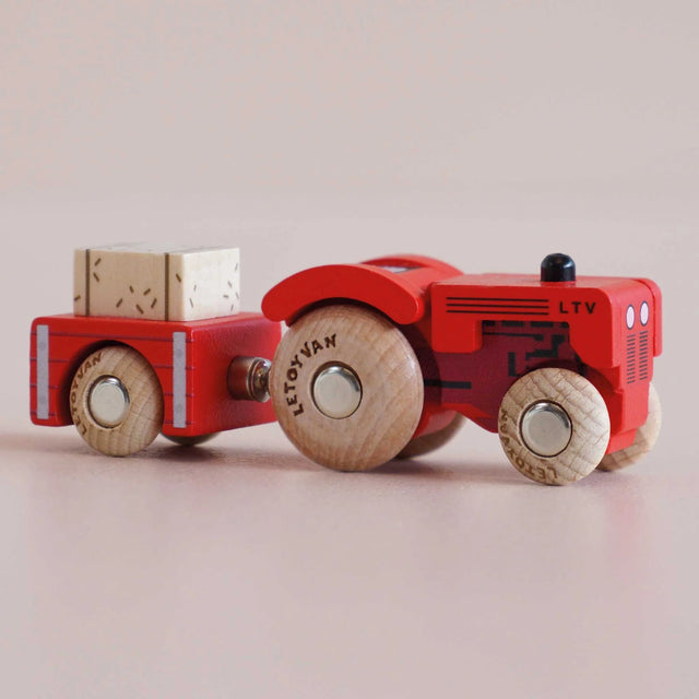Red wooden toy tractor with a matching trailer carrying a hay bale, shown in a side view.