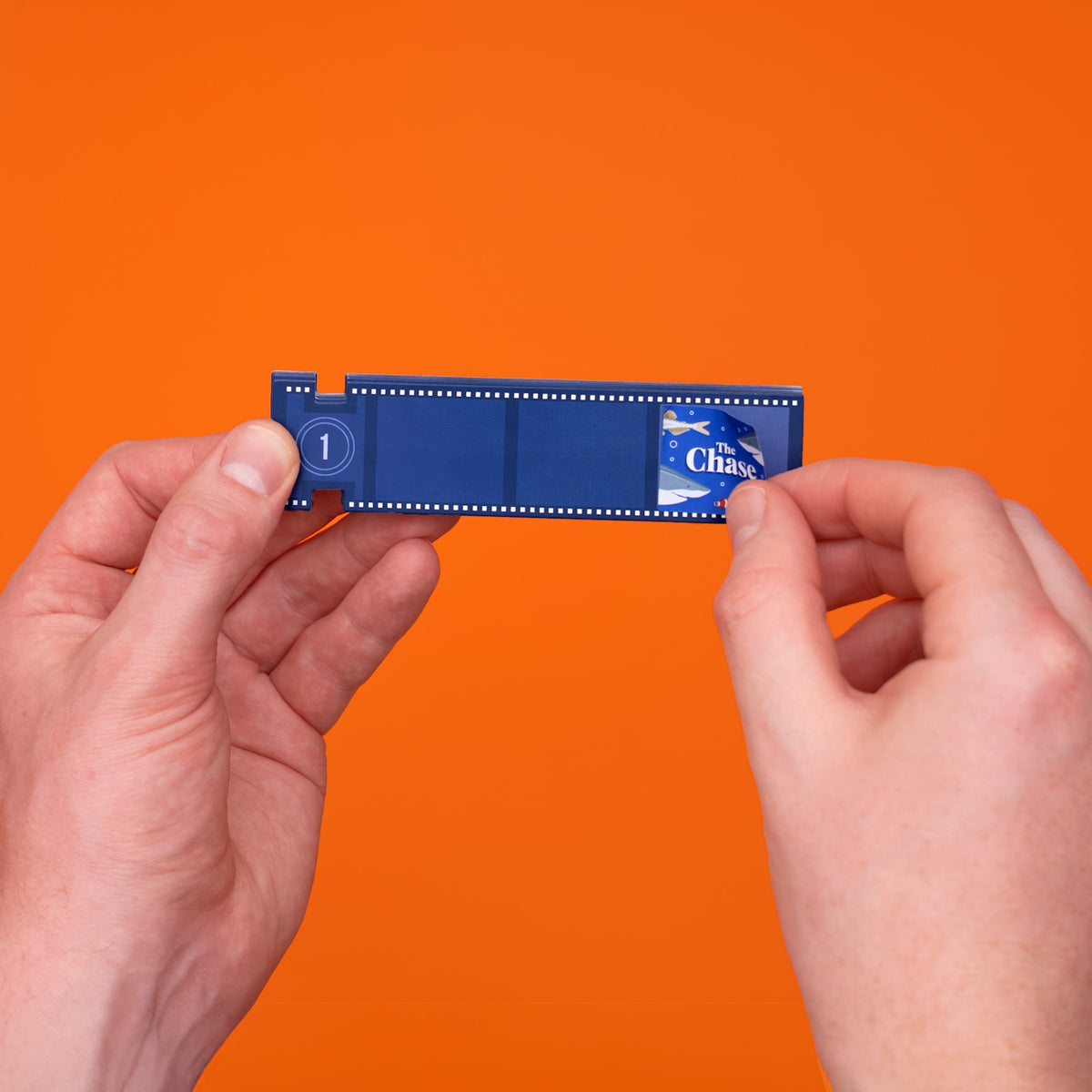 Hands peeling open a blue film strip paper revealing an ocean scene on a bright orange background.