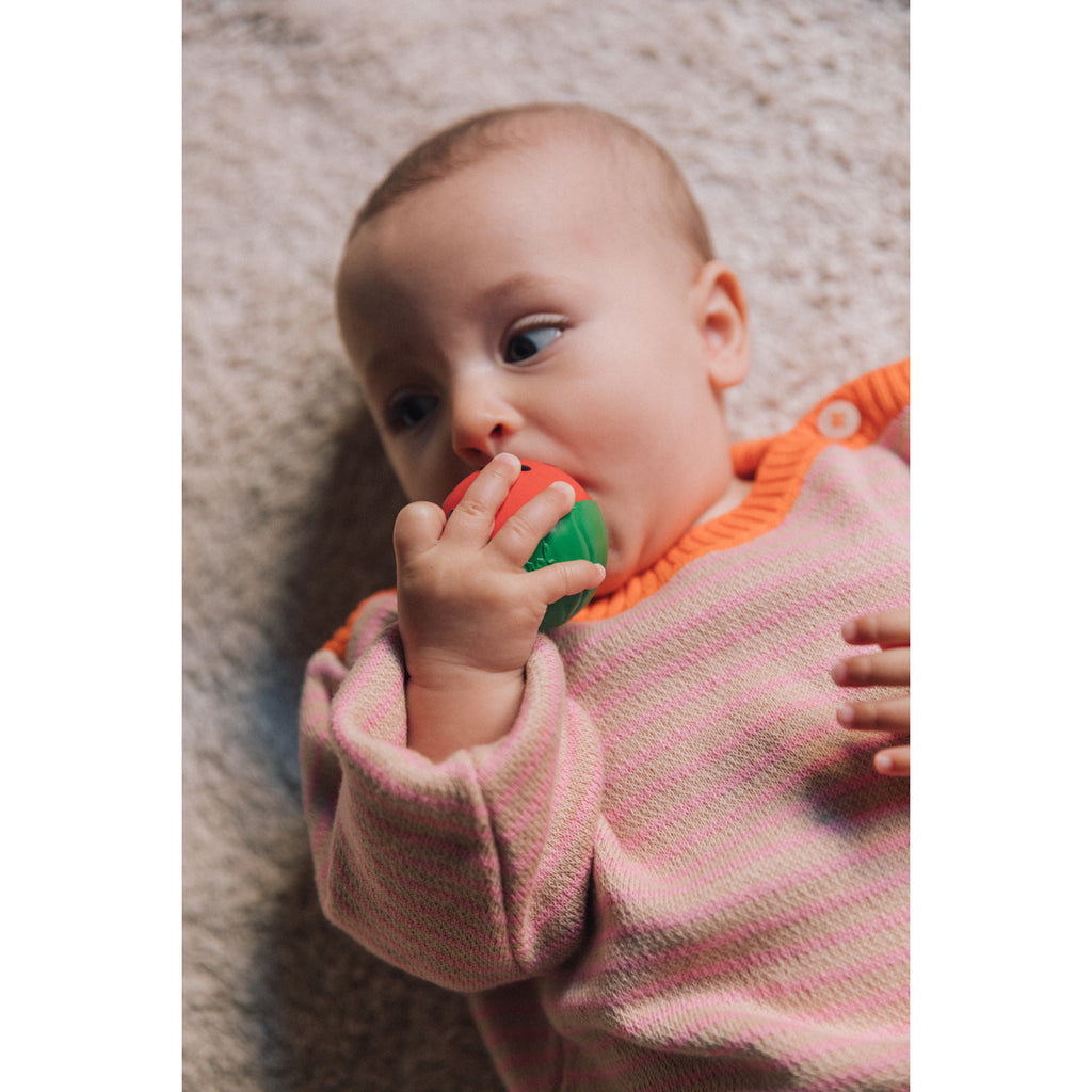 A baby in a pink and orange outfit lies on a soft beige surface, holding and nibbling on a red and green toy, conveying curiosity and playfulness.