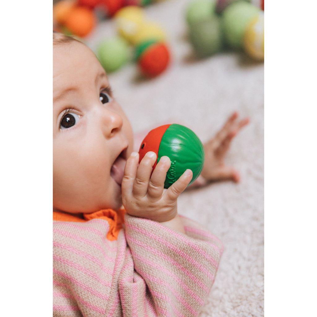 A baby in a pink and orange outfit lies on a soft beige surface, holding and nibbling on a red and green toy, conveying curiosity and playfulness.