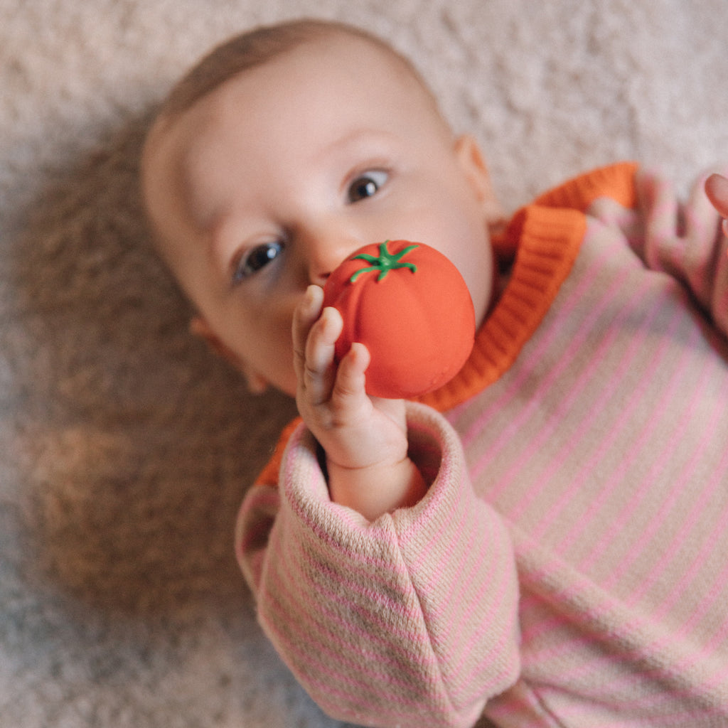 A baby in a pink and orange striped sweater lies on a soft surface, holding a toy tomato to their mouth, conveying curiosity and playfulness.