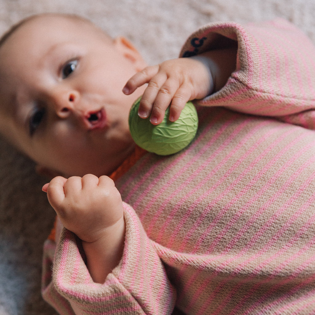 A baby in a pink and white striped outfit lies on a soft surface, holding a small green textured ball. The baby looks curious and engaged.