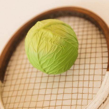 Green cabbage-shaped ball on a vintage wooden tennis racket, with intricate leaf patterns and visible brand lettering, creating a playful, whimsical tone.
