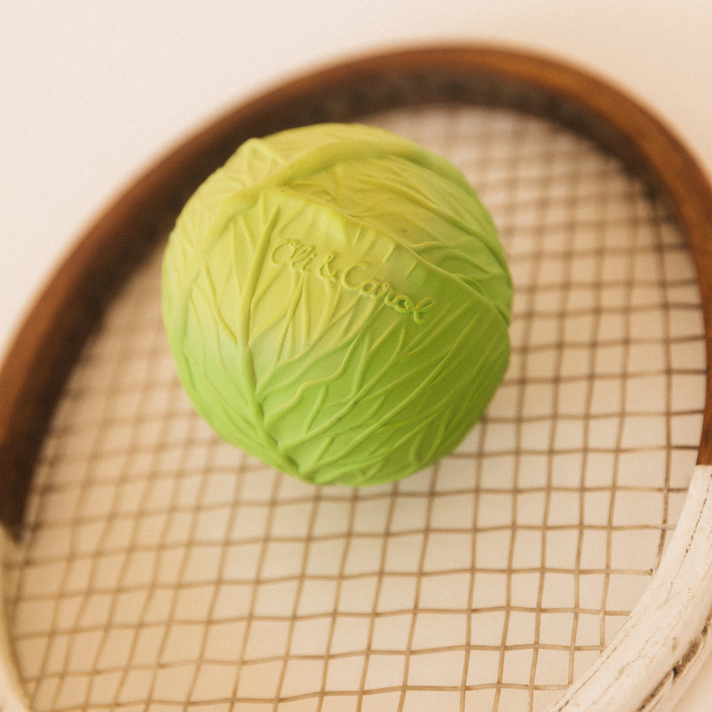 Green cabbage-shaped ball on a vintage wooden tennis racket, with intricate leaf patterns and visible brand lettering, creating a playful, whimsical tone.