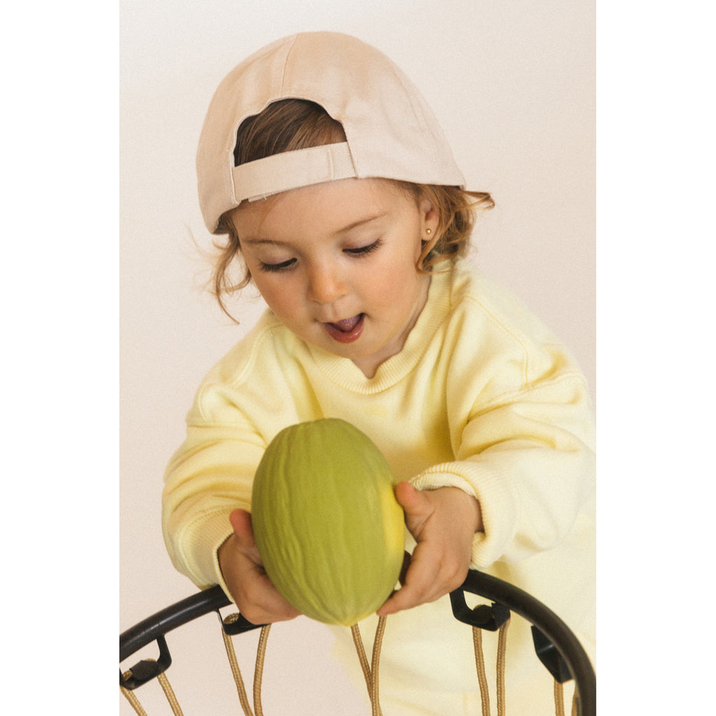 A young child in a yellow sweater and beige cap eagerly examines a green toy fruit while leaning over a black wire basket. The mood is playful and curious.